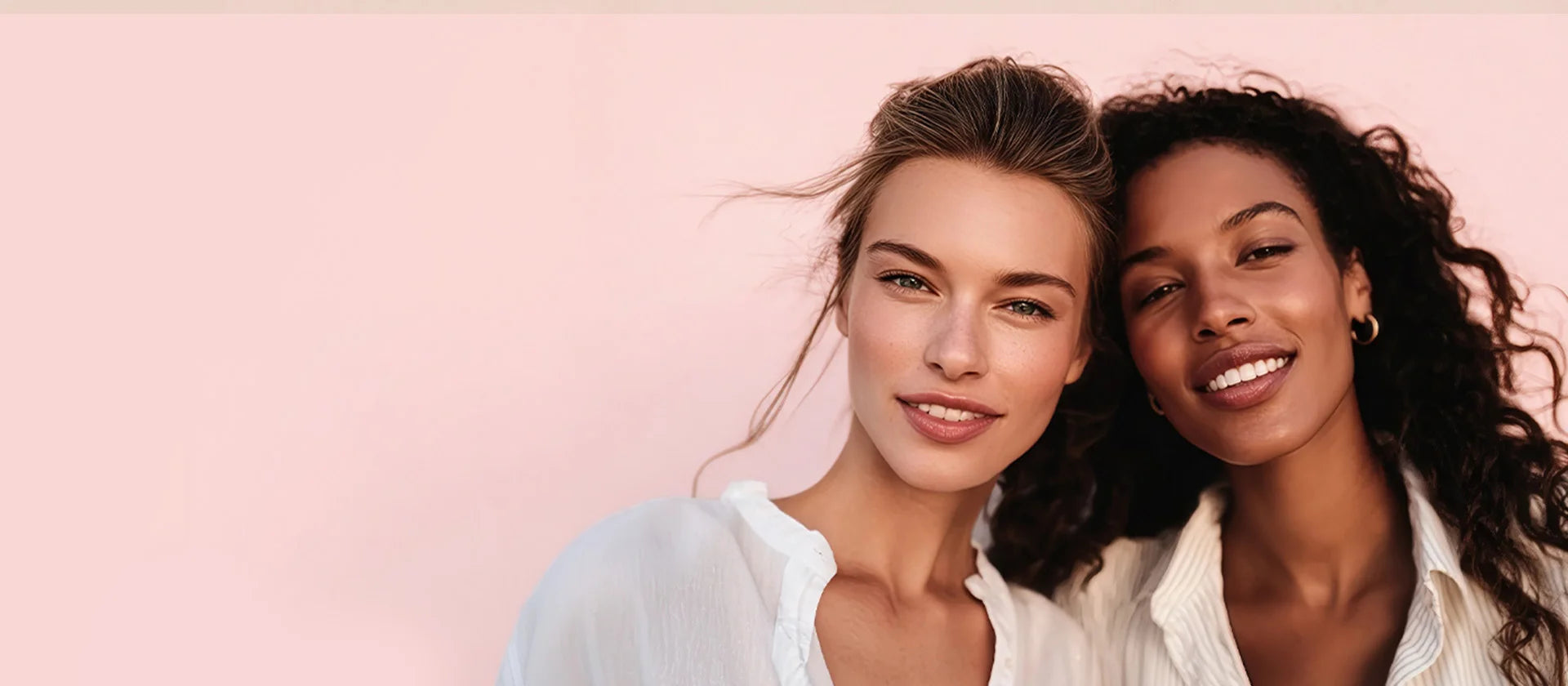 Two smiling women of diverse ethnicities, wearing white tops, posing closely together against a soft pink background