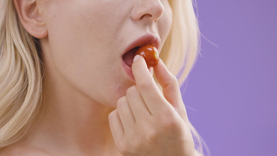 Close-up video still of a blonde woman holding a cherry tomato to her mouth, set against a solid purple background, with a video player interface showing a 0:46 duration