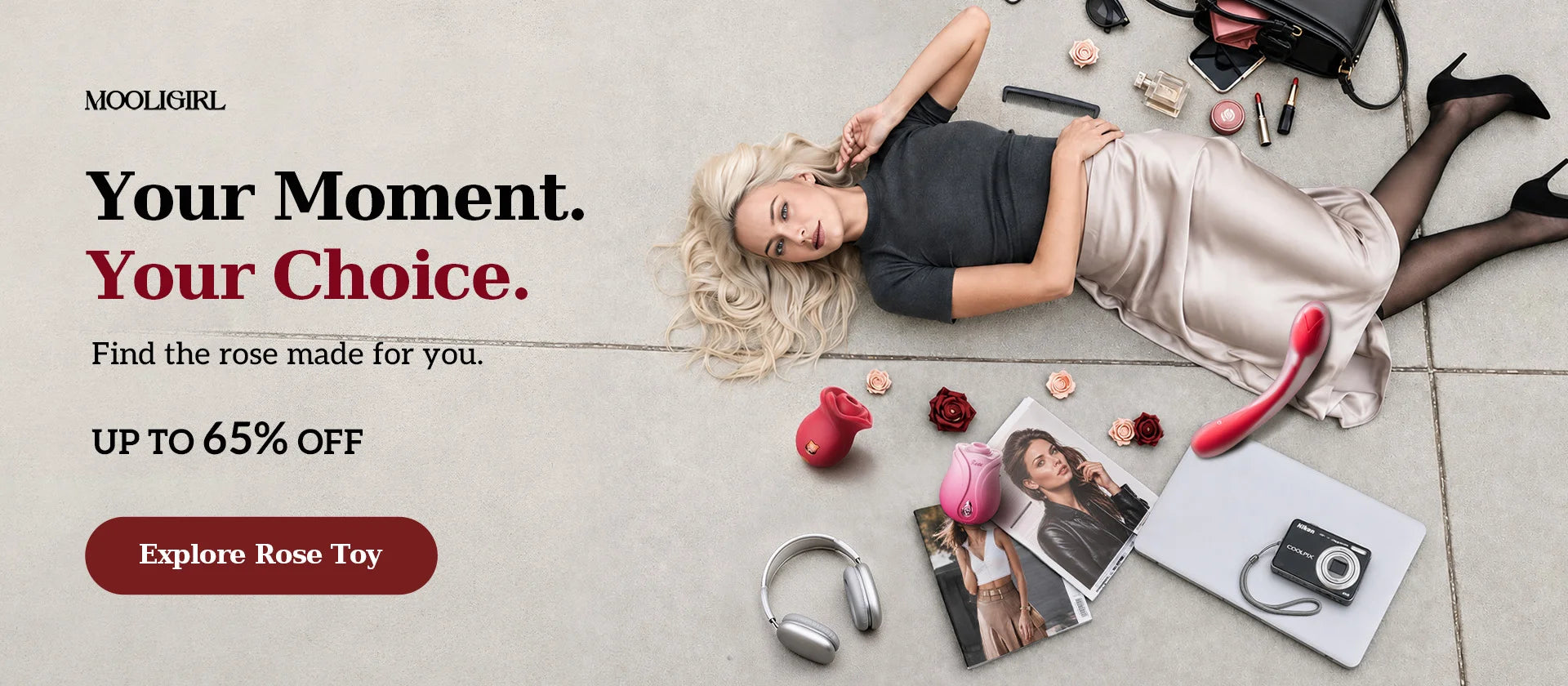 MOOLIGIRL Rose Toy horizontal PC banner, top-down lifestyle shot on light grey concrete floor, blonde woman lying down surrounded by rose-shaped intimate toys, laptop, camera, magazines, headphones and beauty accessories. Left side slogan "Your Moment. Your Choice.", up to 65% off text, burgundy "Explore Rose Toy" CTA button